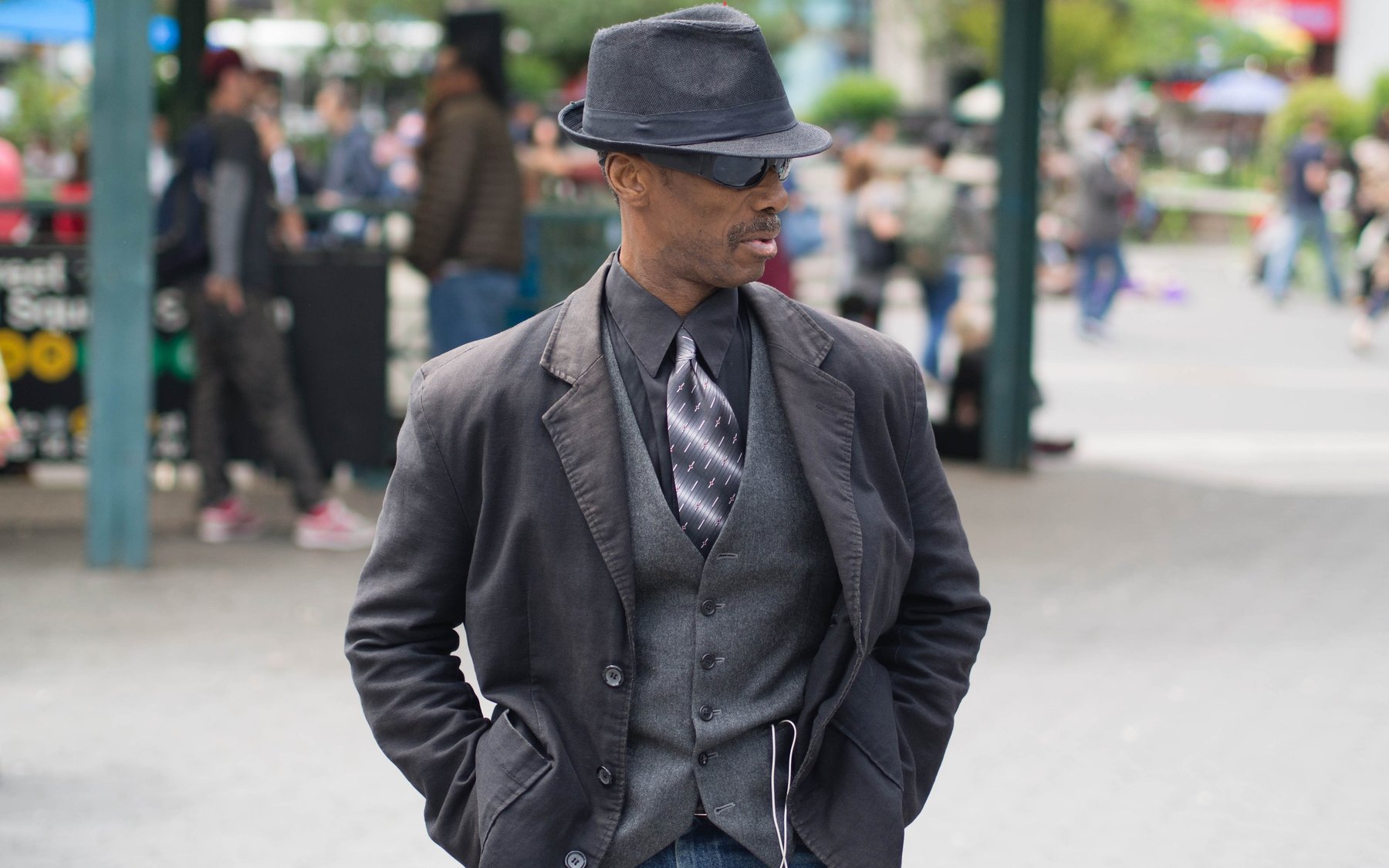 A man in a dark suit and hat standing among a crowd outdoors.