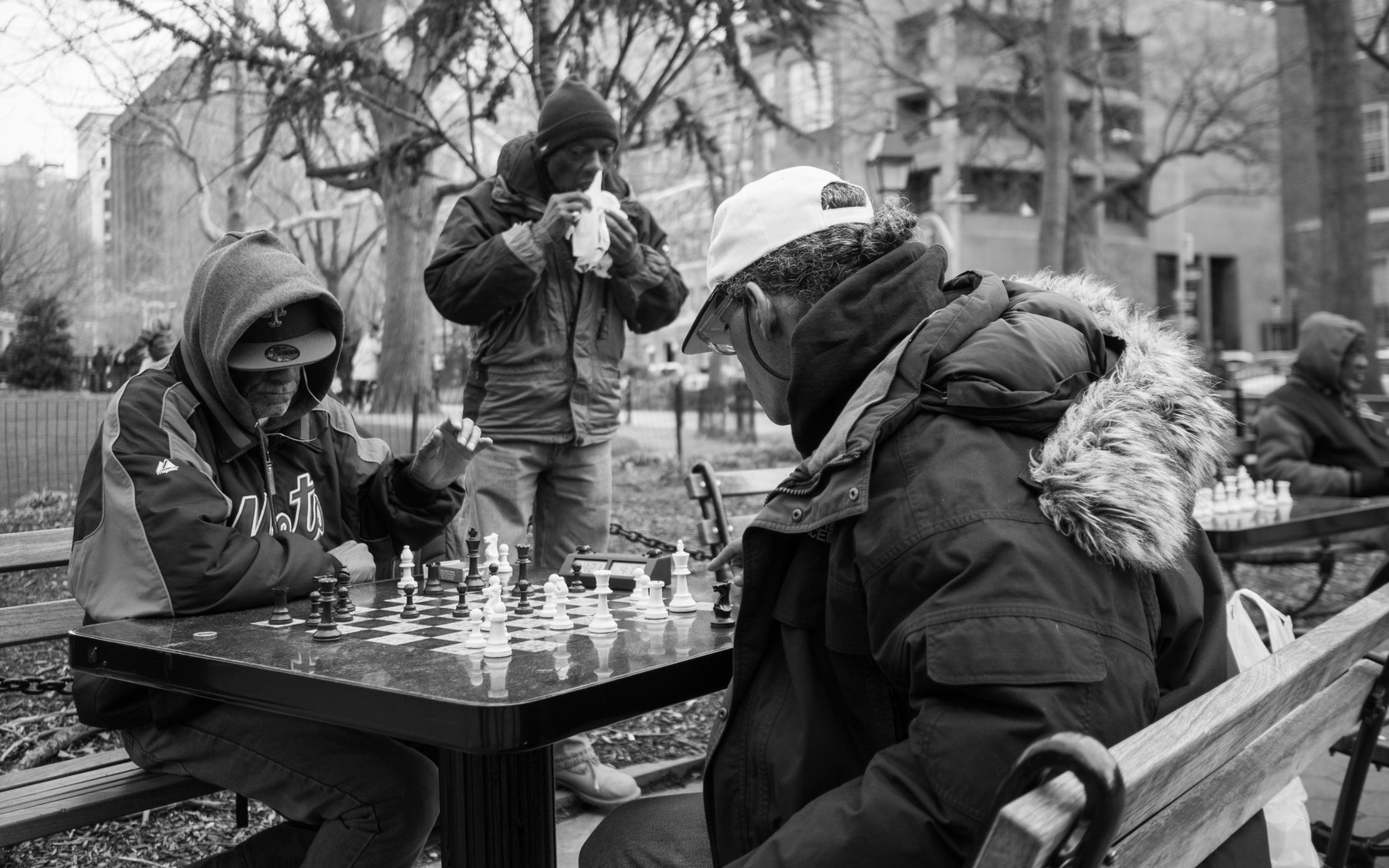 Men leaning over a chessboard beneath bare trees, in black and white.